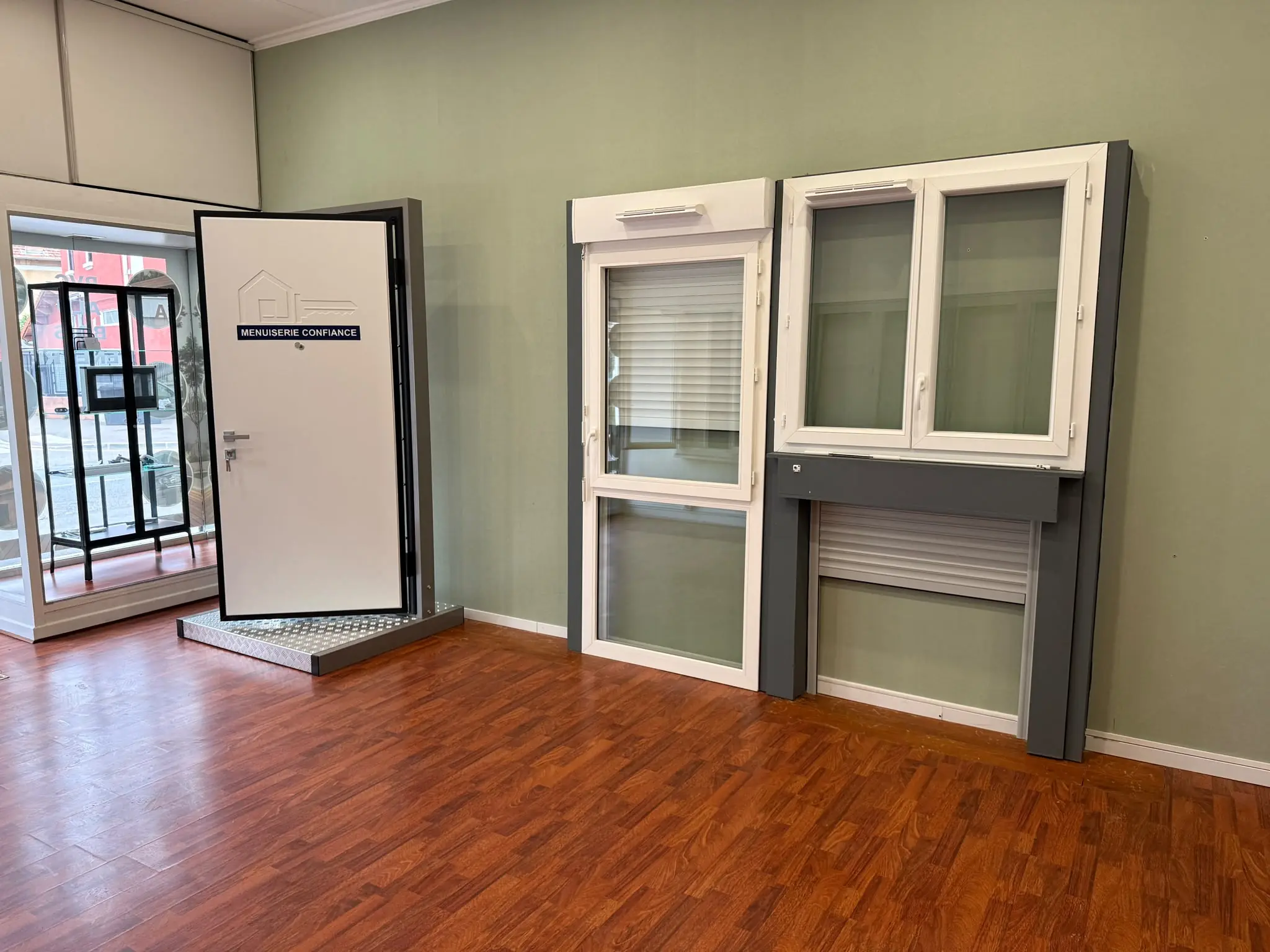 Porte blindée blanche marquée Menuiserie Confiance sur un socle en aluminium, à côté de deux fenêtres blanches et d’un volet roulant exposés contre un mur vert