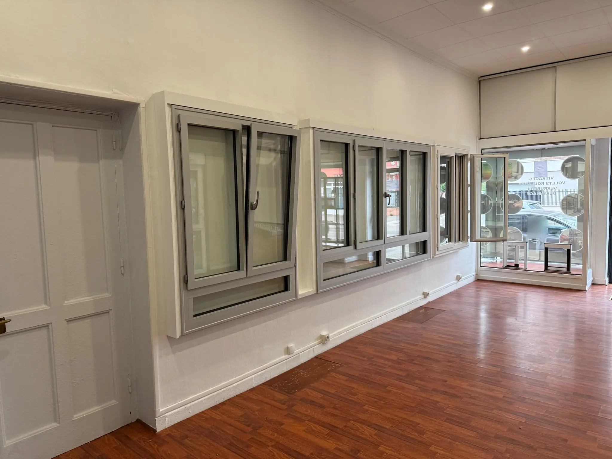 Mur blanc de la boutique où sont exposées plusieurs fenêtres à cadre gris, l’une à deux vantaux au-dessus d’une partie fixe, une autre à quatre vantaux