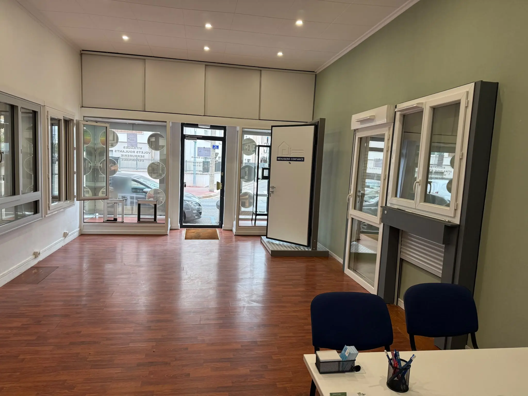 Intérieur de la boutique : murs vert et blanc, fenêtres et porte blindée exposées, entrée vitrée, bureau et deux chaises au premier plan