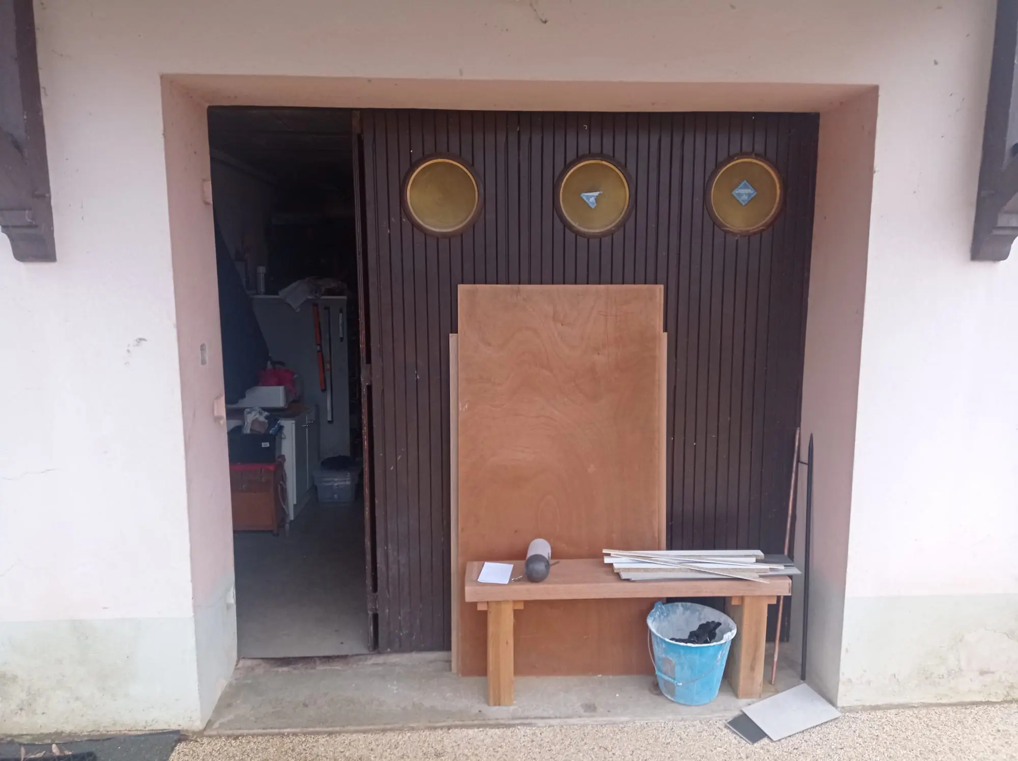 Ancienne porte de garage en bois brun à lames et trois hublots, un vantail ouvert sur le garage