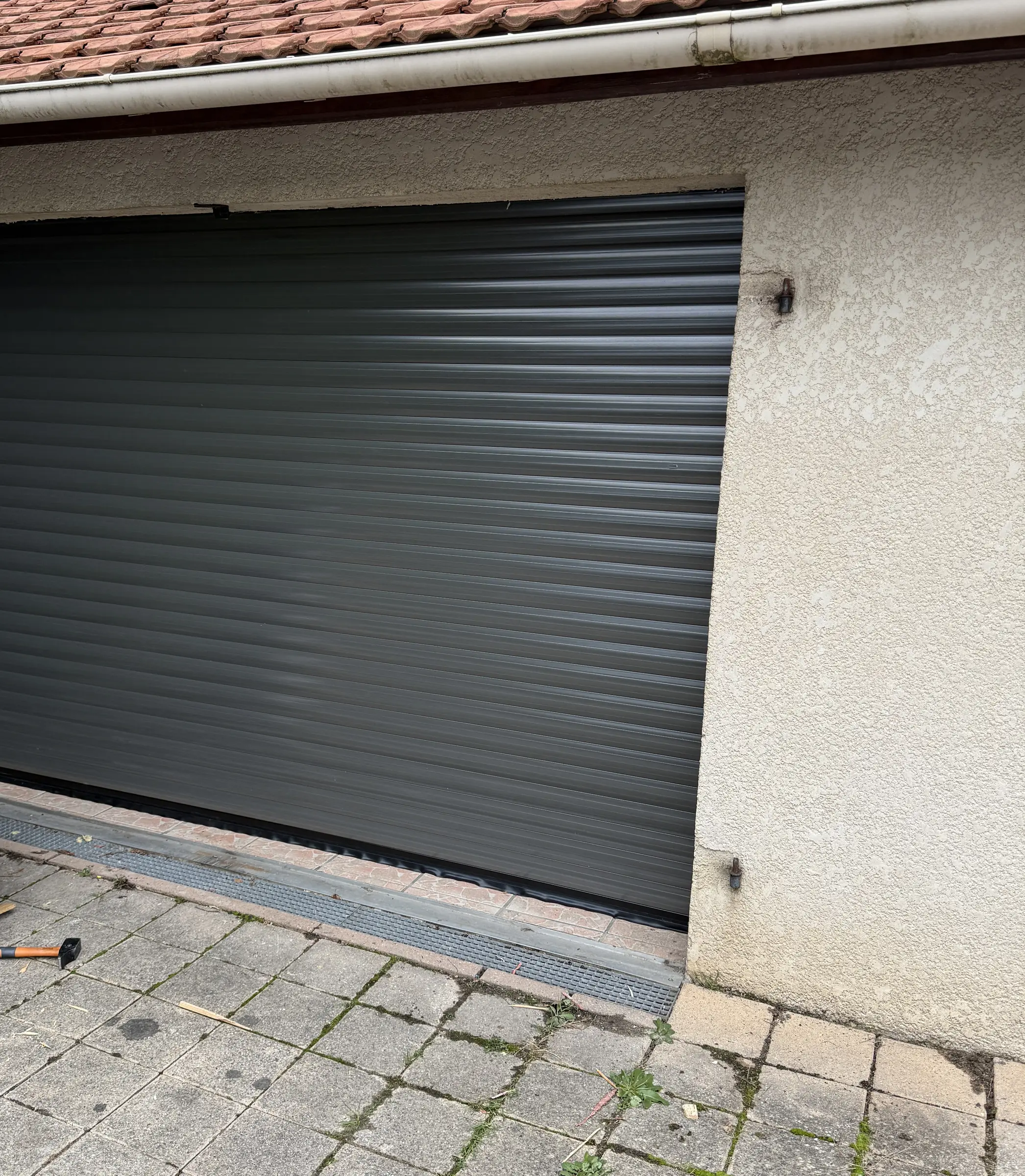 Porte de garage enroulable gris anthracite fermée dans une façade crépie beige, dallage devant